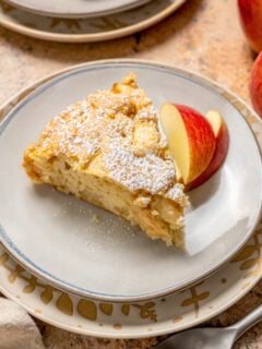 A slice of fluffy apple cake with powdered sugar on top and sliced apples on the side.