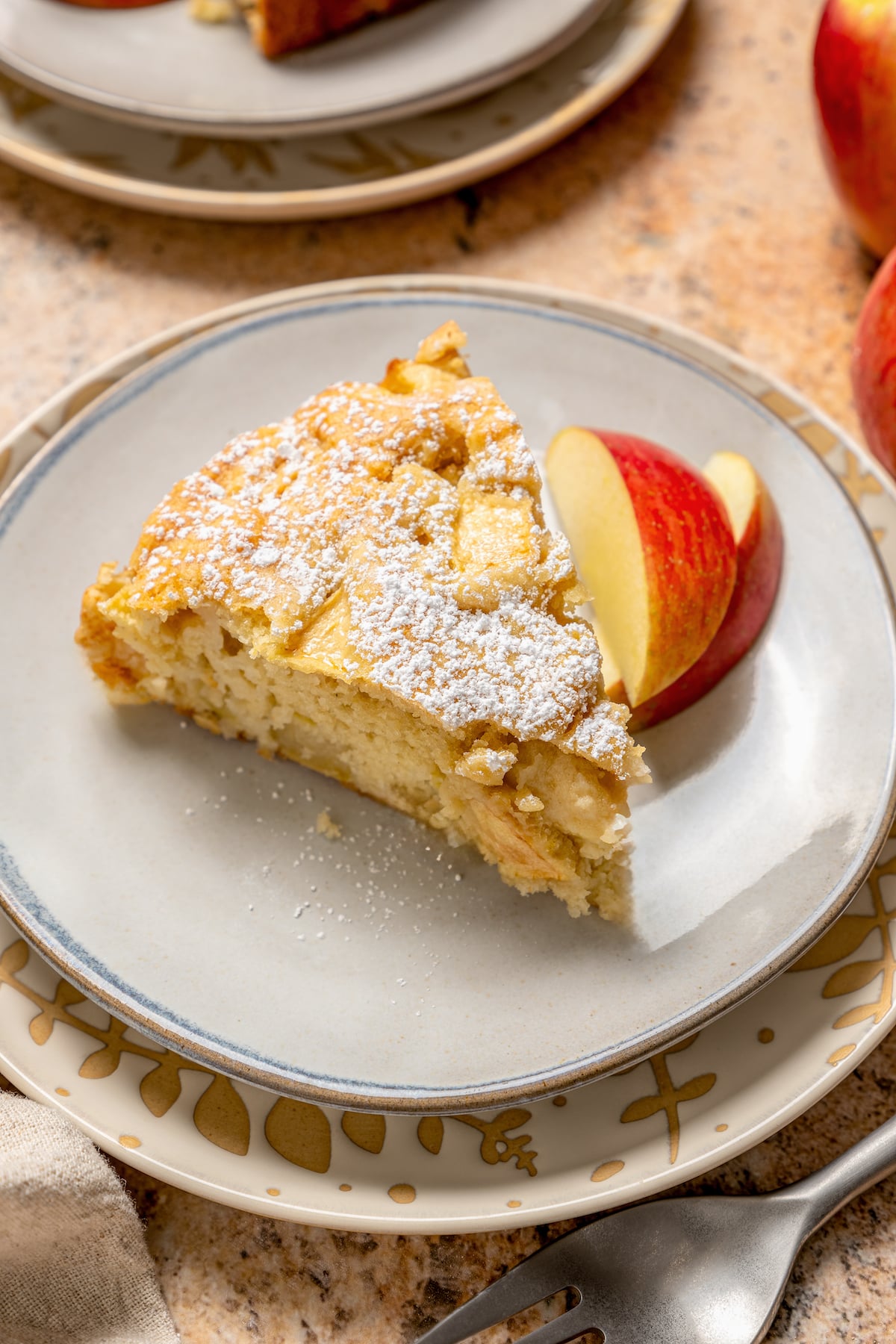 This fluffy apple cake recipe dusted with powdered sugar is an easy fall-flavored dessert once the leaves start falling. A slice of fluffy apple cake with powdered sugar on top and sliced apples on the side.