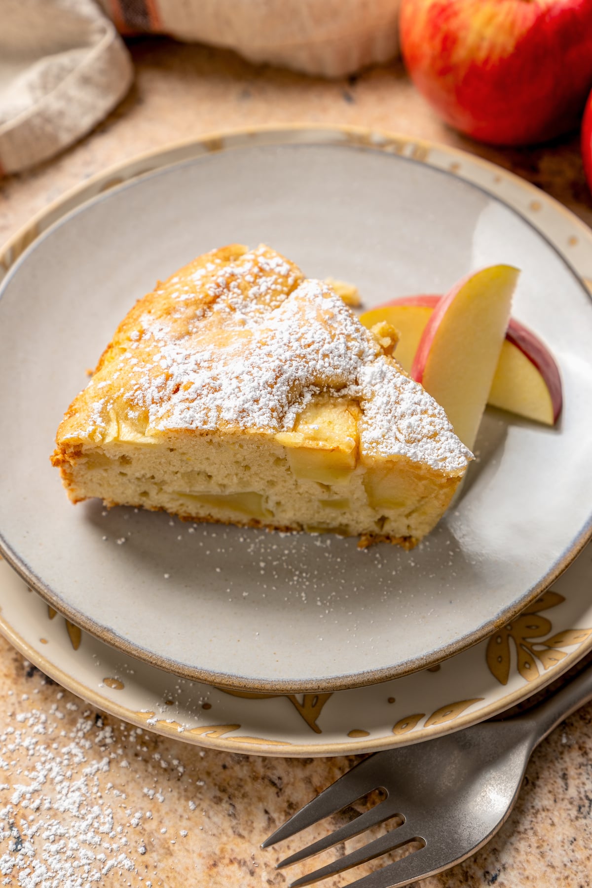 This fluffy apple cake recipe dusted with powdered sugar is an easy fall-flavored dessert once the leaves start falling. Close-up of a slice of fluffy apple cake with powdered sugar.