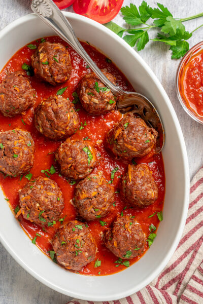 Overhead shot of a white baking dish of meatballs with a striped cloth napkin, herbs, and sauce.