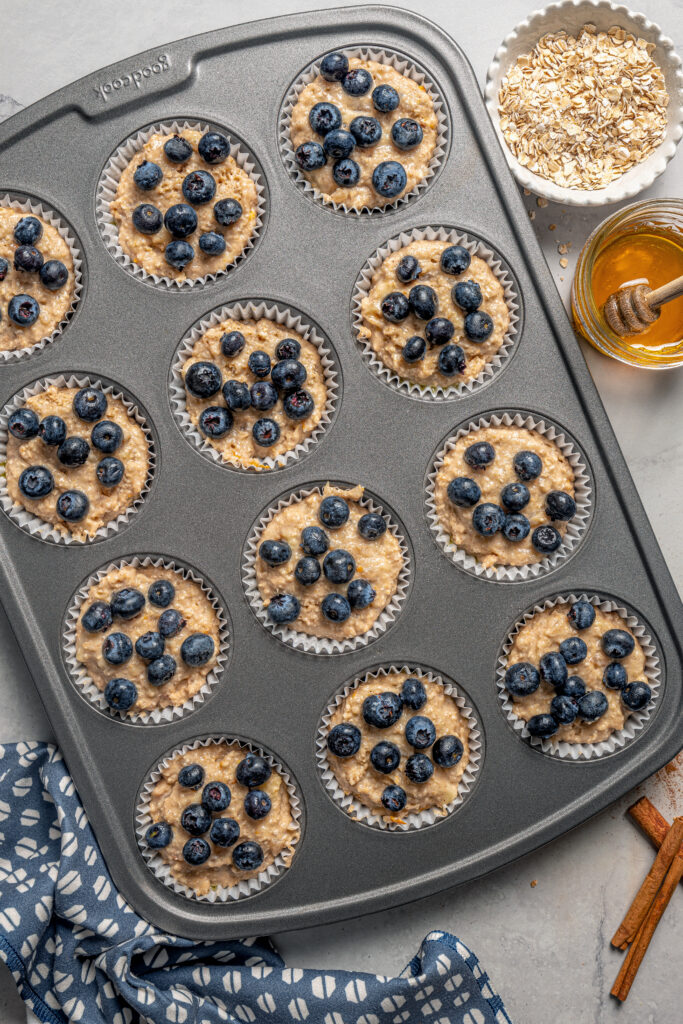 This naturally-sweetened blueberry protein muffins recipe with cinnamon and orange zest is a great on-the-go breakfast. Liners filled with batter and topped with fresh blueberries.