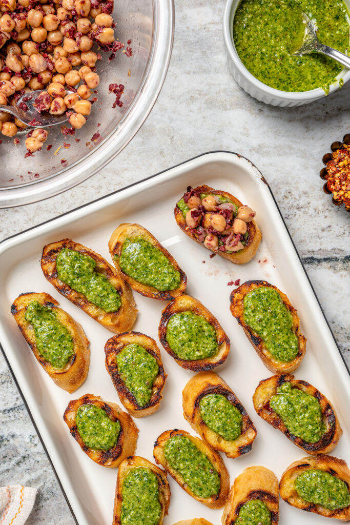 This zesty chickpea bruschetta recipe with herby pesto and salty olives on toasty bread is a 20-minute summer appetizer. Assembling the appetizers with pesto and the chickpea mixture.