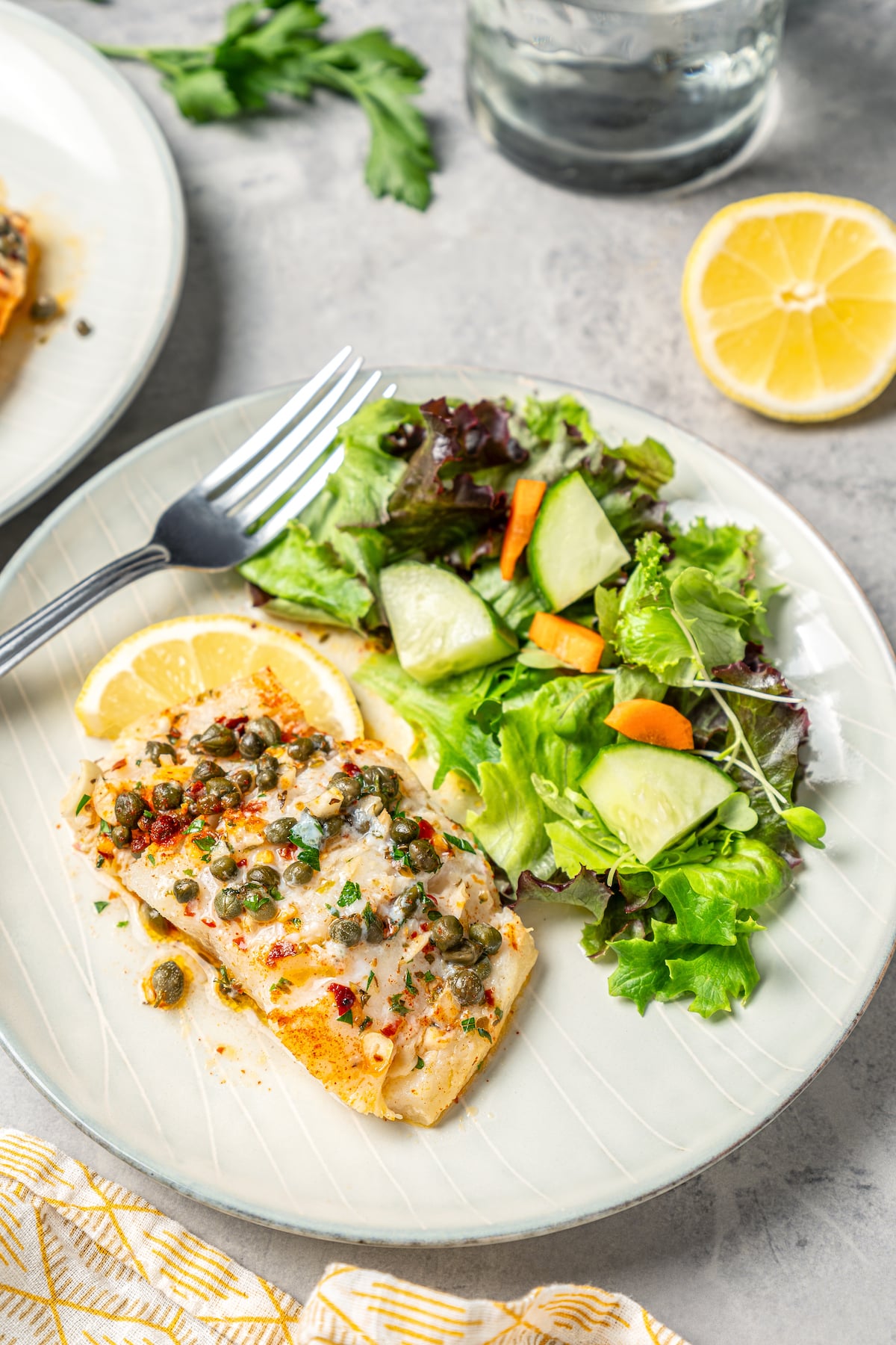 You'll crave the light, fresh flavors of this easy Baked Cod Recipe. Simple prep, perfect texture, and zingy flavor - what could be better? A dinner plate with a portion of fish and a side salad.