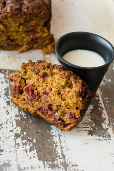 A slice of cranberry loaf cake next to a cup of milk. The remainder of the loaf is on the table in the background.