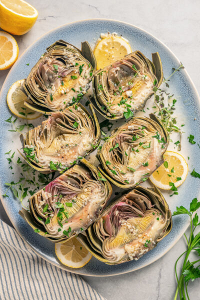 Halved artichokes with garlic, fresh herbs, and lemon wedges.