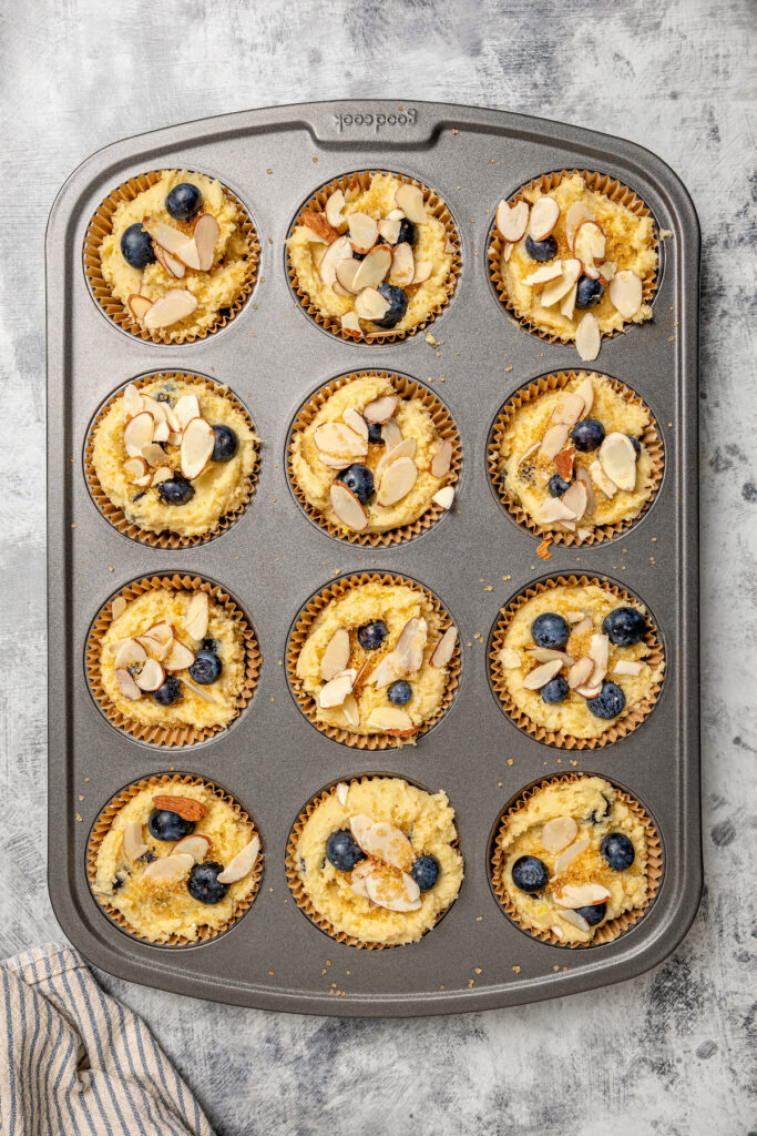 These lemon-blueberry ricotta muffins with crunchy almonds are an easy 20-minute baked treat. They're so light and fluffy you'd never guess they're gluten-free! Add shredded coconut for extra fruity vibes. Liners filled with batter and topped with almonds.