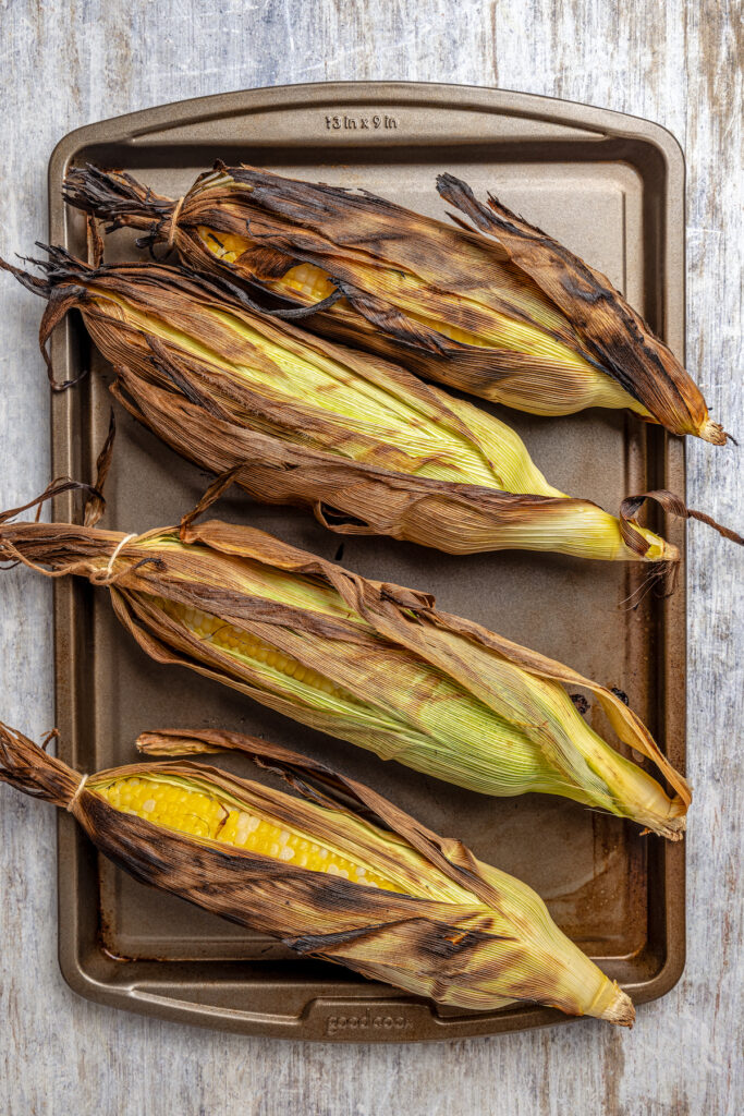Sweet, fresh corn is a must-have in the summer, and this easy Grilled Corn is loaded with a unique nutty taste that you're sure to love! Grilled corn in the husk on a baking sheet.