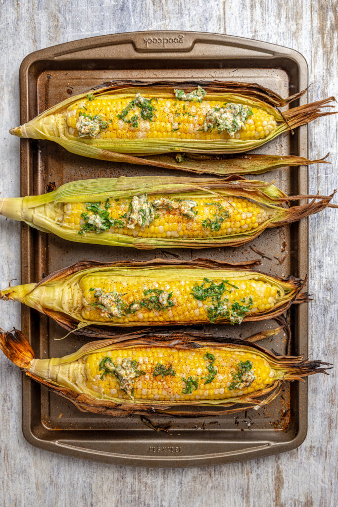 Sweet, fresh corn is a must-have in the summer, and this easy Grilled Corn is loaded with a unique nutty taste that you're sure to love! Ears of grilled corn with part of the husk peeled away, showing the kernels. Seasoned butter has been dabbed on top.