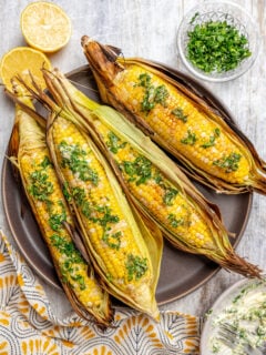 A plate of corn cooked in the husk, next to dishes of toppings.