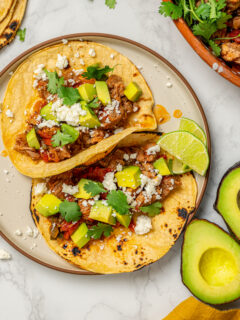 Two pulled pork tacos on a plate with queso fresco, cilantro, and avocado.