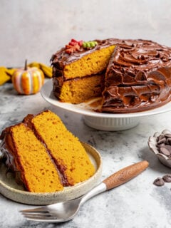A slice of pumpkin cake with chocolate frosting on a serving plate, and the rest of the cake in the back.