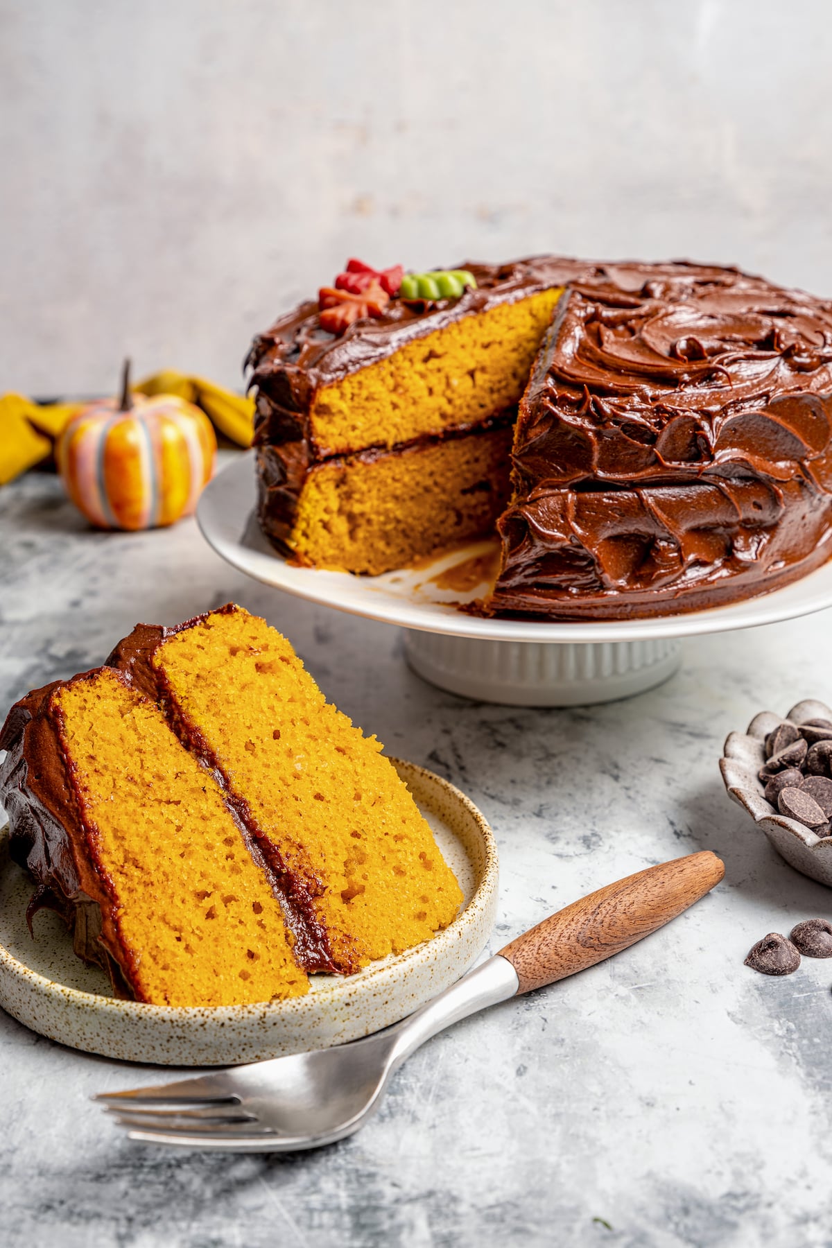 This warmly spiced pumpkin cake recipe with velvety cream cheese-chocolate frosting is a sweet end to any Thanksgiving dinner. A slice of pumpkin cake with chocolate frosting on a serving plate, and the rest of the cake in the back.