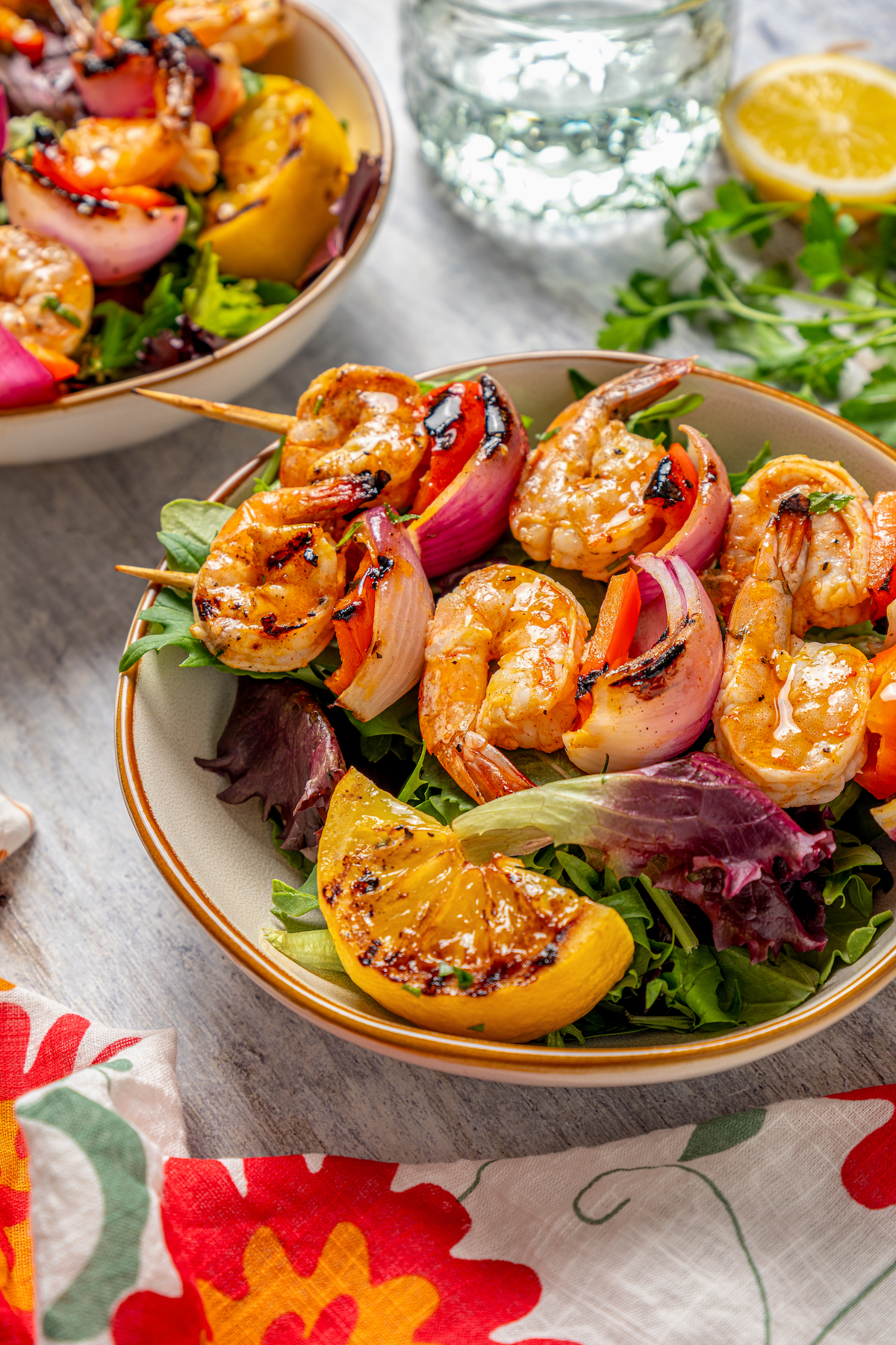 This shrimp brochette recipe with garlic-lemon marinade, sweet bell peppers, and spicy red onions is an easy 20-minute lunch. Bowl of salad with shrimp skewers and a grilled lemon.