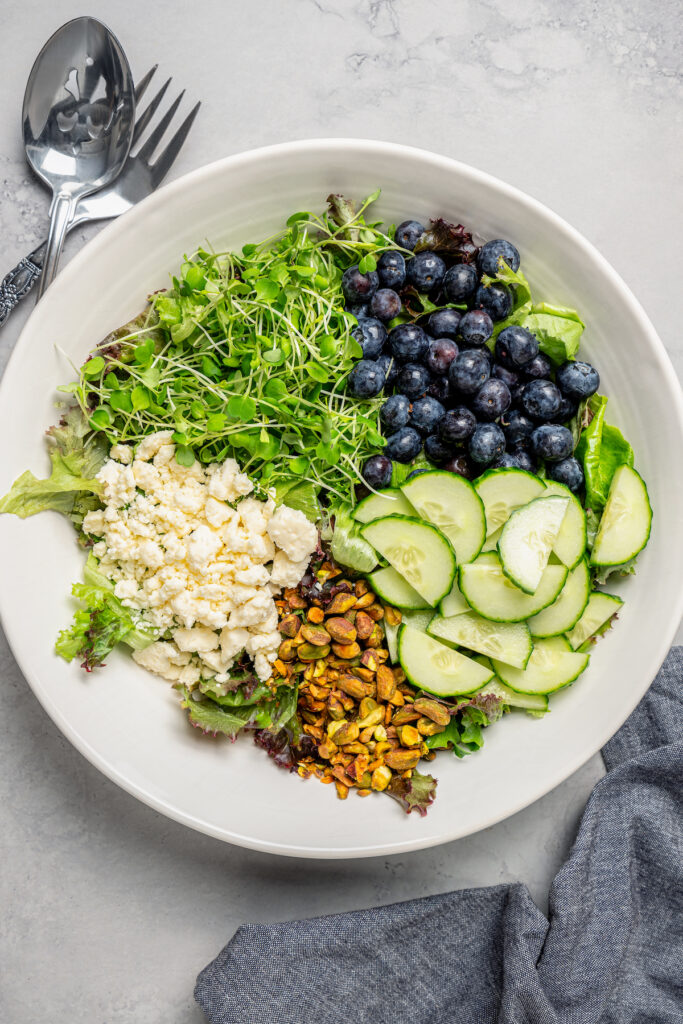 Spring Mix Salad with Champagne Dressing is elegant, flavorful, and unique - and only takes minutes to prepare! A nutritious, paleo salad. A salad bowl with blueberries, microgreens, cucumbers, pistachios, and feta cheese.