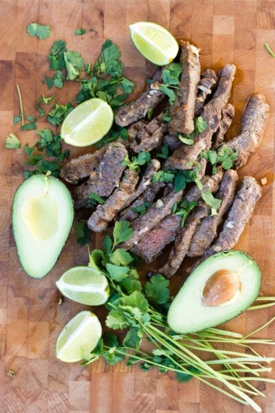 Overhead shot of Cuban mojo steak on a wooden board with avocado halves, limes, and cilantro.