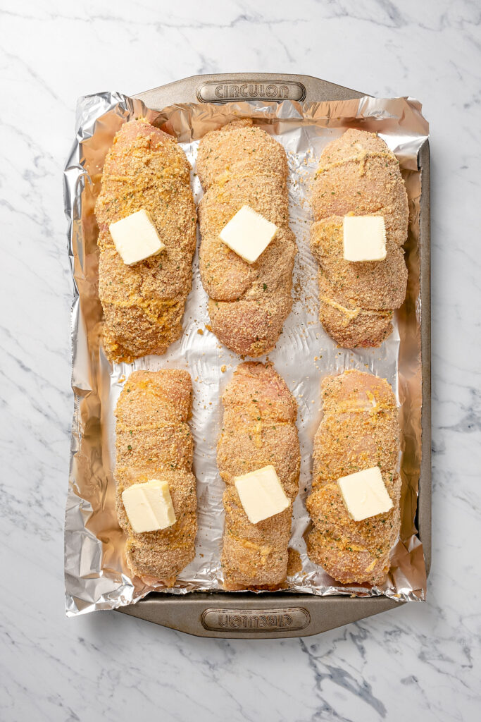 Stuffed Chicken Breasts pack so much flavor in every bite! With goat cheese, basil and white wine mushroom sauce, it's fancy but easy to make. Breaded, stuffed chicken breasts on a baking sheet. Each piece is topped with a pat of butter.