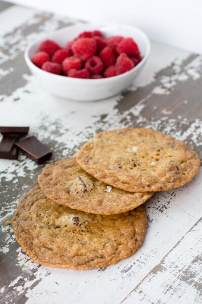 Three thin cookies on a table with a bowl of raspberries behind them